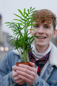 The guy has a nasty smile with braces, looks at the camera, holds a green home pot with a flower 