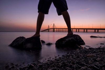 Low section of man standing on rock by sea against sky during sunset