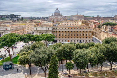 Trees and buildings in city against cloudy sky