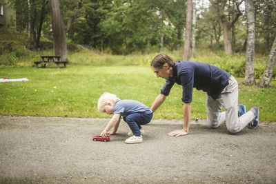 Father kneeling while son playing with toy car on footpath against plants at park