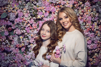 Mother and daughter in white  sweaters stand against a pink floral wall in spring. people smile 