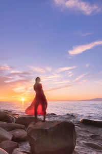 Man sitting on rock by sea against sky during sunset