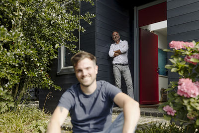 Smiling father leaning on house wall while son sitting in backyard