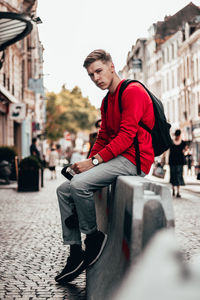 Young man with backpack sitting on barricade amidst street in city