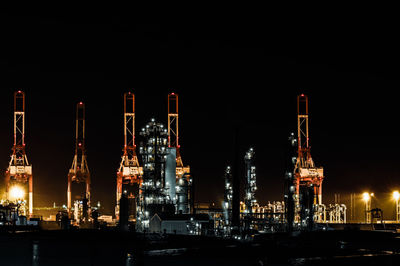 Illuminated factory by buildings against sky at night