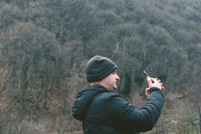 Side view of man photographing against bare trees on mountain