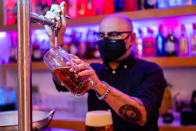 Side view of serious barkeeper in eyeglasses pouring beer into glass while working in bar during coronavirus pandemic