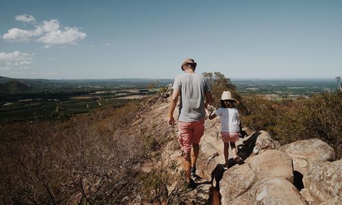 Rear view of father and daughter walking on rocks at mountain against sky