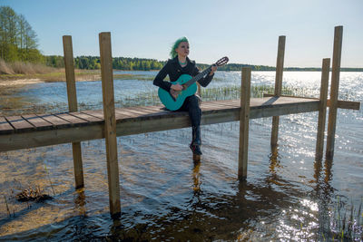 Hipster young woman with turquoise guitar sitting on pier over lake