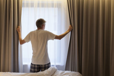 Rear view of man holding curtains while standing at home