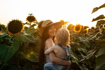 Beautiful mother holds a little son in her arms in a field of sunflowers at sunset