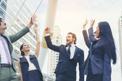 Happy young colleagues with arms raised standing against buildings in city