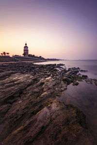 Lighthouse amidst sea and buildings against sky during sunset