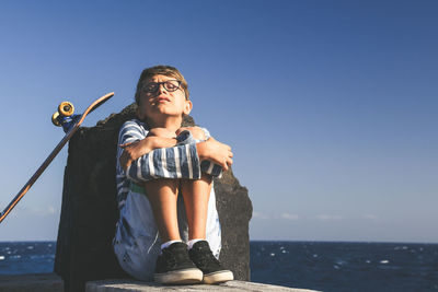 Boy sitting with skateboard by sea against clear sky