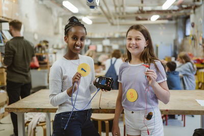 Portrait of smiling female students showing number ranks while holding electrical parts near table in classroom
