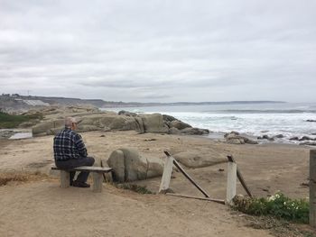 Rear view of man sitting on bench while looking at sea against sky