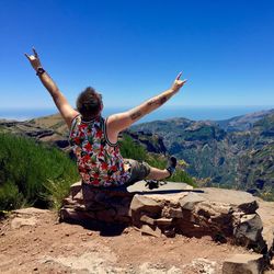 Rear view of man with arms raised sitting on cliff during sunny day