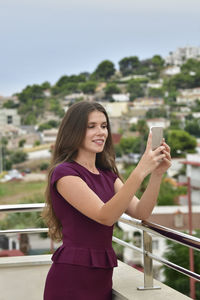 Smiling young woman using mobile phone while standing by railing