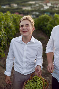 Teenage boy in a white shirt stands in a vineyard at sunset and holds a basket of green grapes