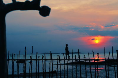 Silhouette people standing on wooden post by sea against sky during sunset