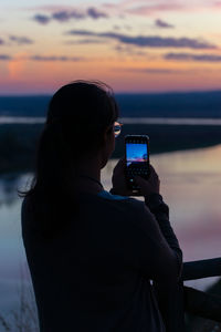 Rear view of woman photographing against sky during sunset