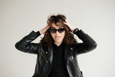 Brunette with curly hair, glasses and leather jacket shows her tongue isolated