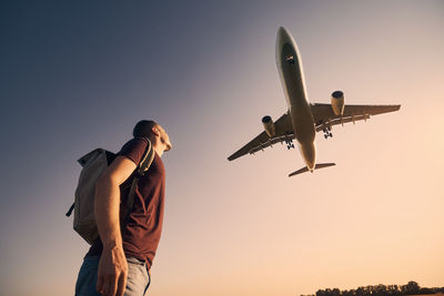 Man with backpack looking up to airplane landing at airport during beautiful summer sunset.