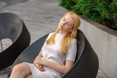 Young beautiful blond woman sitting outdoors, on the wooden terrace in the city and resting. 
