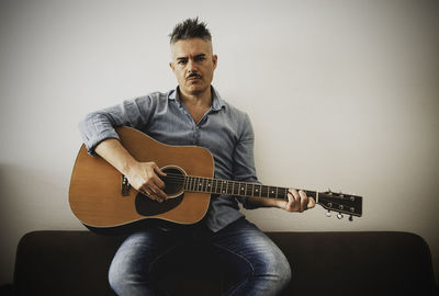 Portrait of man playing guitar while sitting on sofa against wall