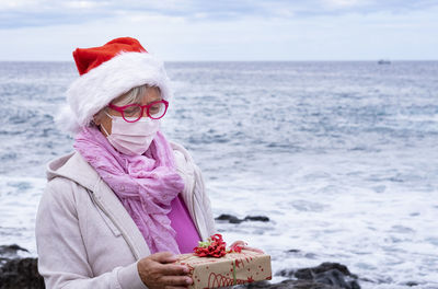 Senior woman wearing mask holding gift standing against sea