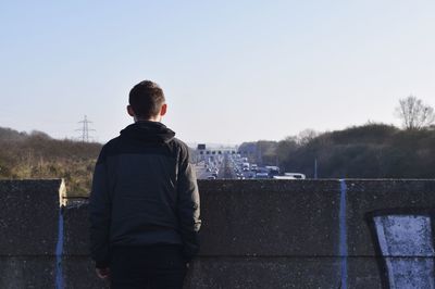 Rear view of man standing by retaining wall against clear sky