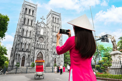 Rear view of woman wearing graduation gown standing against building