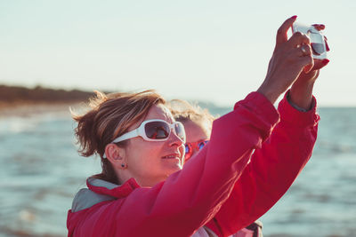 Side view of mother photographing sea while standing with daughter against sky during sunset