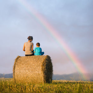 Rear view of mother and son sitting on hay bale against sky during sunset