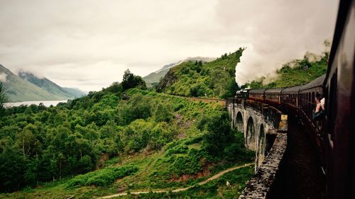 Train on bridge leading towards green mountain against cloudy sky