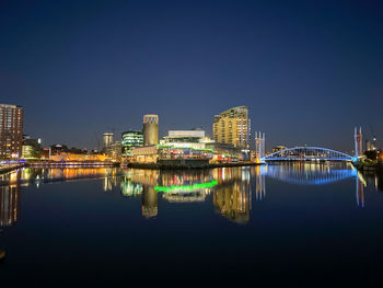 Illuminated buildings by river against clear blue sky at night