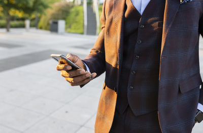 Unrecognizable ethnic businessman in stylish suit standing in city and surfing internet on smartphone while working remotely on project