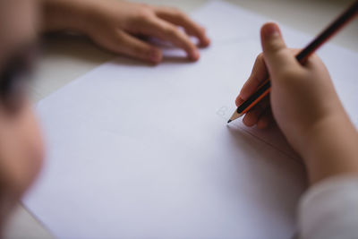 Crop cute kid sitting at wooden table and drawing with pencil on paper while relaxing at weekend at home