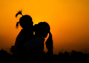 Silhouette mother carrying daughter while standing against orange sky