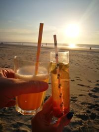 Close-up of hand holding drink at beach against sky during sunset