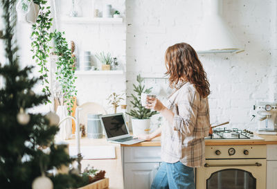 Attractive smiling woman with curly hair in plaid shirt with laptop near window at bright kitchen 