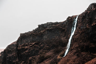 Low angle view of rocky mountains with small waterfall against cloudy sky