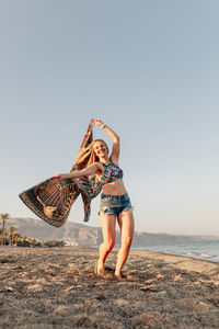 Full length of happy woman with scarf standing at beach against clear sky