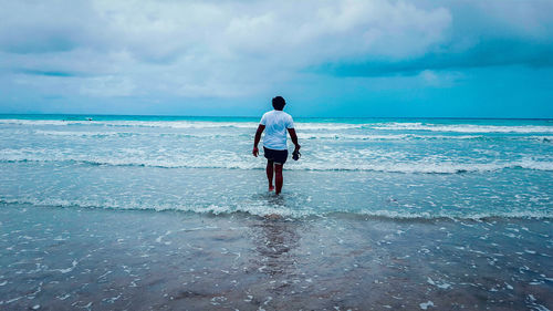 Rear view of man walking on shore at beach against cloudy sky