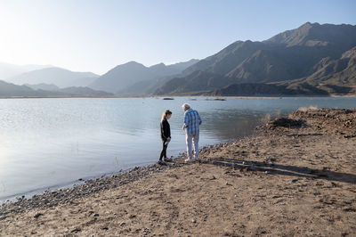 Senior man walking with his granddaughter along the lake shore with mountains.