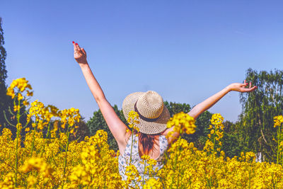 Rear view of woman with arms raised standing amidst flowering plants against clear blue sky
