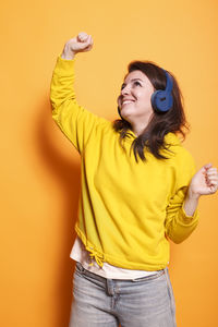 Young woman with arms raised standing against yellow background