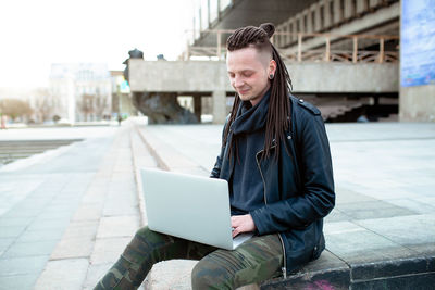 Hipster man using laptop on footpath in city