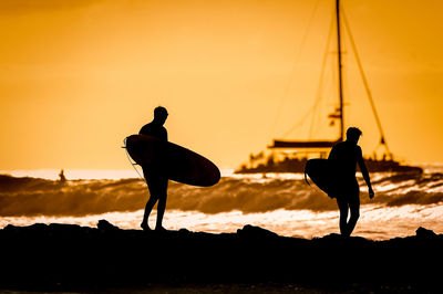 Silhouette teenage boys with surfboards standing at beach against clear sky during sunset