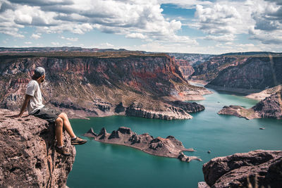 Man looking at view while sitting on cliff against mountains and cloudy sky